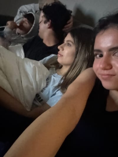 The image shows a group of four people in an indoor setting. A person with dark hair is taking a selfie, their face dominating the upper portion of the frame. Below them is a person with long brown hair, wearing a light blue t-shirt and seemingly covered with a white blanket. To the left are two other individuals, partially obscured by the group, one of whom is wearing a grey hooded sweatshirt. The overall lighting is dim, creating a somewhat muted atmosphere. Based on the location in the city of College Station, United States of America, it could be a dorm room, an apartment or a house. The subjects appear to be in a close social gathering. There is no readily discernible specific address.