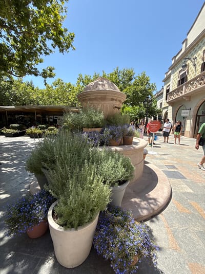 The image shows a sun-drenched, pedestrianized street in La Roca del Vallès, Spain, likely within the La Roca Village shopping outlet, characterized by its distinct architecture and outdoor mall layout. In the foreground, a large, multi-tiered stone fountain is decorated with abundant potted plants, including tall, leafy green shrubs and smaller plants with delicate blue flowers. The paving stones are laid in a geometric pattern with some reddish-brown inlays.

In the mid-ground, people are walking along the street. To the left, there's a covered outdoor seating area for a restaurant or cafe, with umbrellas visible and patrons seated at tables. Further down the street, various shops are visible, including one with a prominent "LOEWE" sign. The architecture of the buildings features decorative facades with patterned elements and balconies.

A white, circular sign with text in orange and black stands on the right side of the fountain, partially obscured by a person wearing a red t-shirt. Several individuals are browsing or walking, dressed in casual summer attire. The overall environment suggests a pleasant, bright, and warm day, with clear blue skies and dappled sunlight casting shadows on the ground from overhead trees and structures.