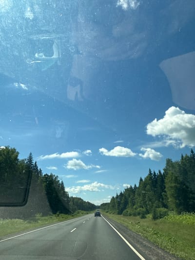 The image depicts a view from inside a vehicle traveling on a paved road lined with dense green trees on both sides. The sky above is a clear blue with scattered white clouds. A dark-colored car is visible in the distance on the same road, heading in the same direction. The foreground shows the dashboard of the vehicle and its windshield, which has several smudges and specks. The scene appears to be a highway or a well-maintained road through a forested area, likely in a rural or suburban setting. There are no visible people or specific landmarks to indicate the exact location within the city of Nikolskoye, Russia, other than the general environment.
