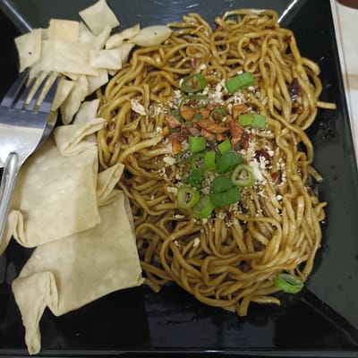 A close-up shot of a plate of Mie Goreng (fried noodles), commonly found in Cangkring, Indonesia, is shown. The noodles, topped with chopped green onions and fried shallots, are situated on a black square plate. To the left of the noodles is a pile of fried wonton wrappers, with a fork laying on top of them. The background is out of focus.