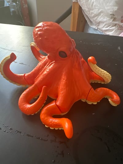 An orange plastic octopus toy is positioned on a dark, textured surface, possibly a table, with a few scattered water droplets and specks of dust visible. The octopus has a bulbous head with two dark, inset eyes. Its eight arms, lined with lighter colored suction cups, are spread out in various positions, some curled and others extended. The toy appears to be segmented, with visible lines along its arms and body. In the background, a wooden headboard with a white patterned duvet is visible, along with blurry objects that suggest an indoor environment. The lighting in the scene is bright, casting reflections on the toy's surface. The image does not provide any information about its location in Hradec Králové, Czechia.