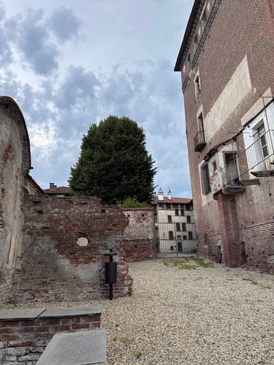 In Lagnasco, Italy, a wide-angle, low-angle shot captures a courtyard with a gravel ground. To the right, a tall, imposing brick building with arched decorative elements at the top dominates the frame, its rough texture evident. A small, dilapidated balcony with an open doorway and rusted metal railing juts out from its facade, revealing a window behind it. To the left, a crumbling brick wall, showing signs of disrepair and wear, forms a stark contrast. In the background, a large, dense green tree stands prominently. Beyond the tree and to the left, other buildings with tiled roofs and classical architectural elements, including two distinct white spires, are visible. A metal gate separates the courtyard from these background structures. A black trash receptacle on a stand is positioned near the broken brick wall. The sky is overcast with scattered clouds.