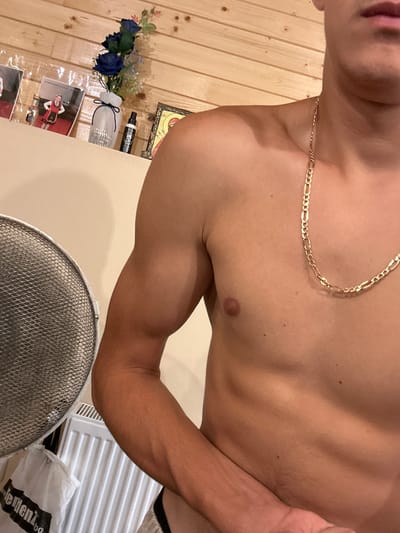 A shirtless person is shown from the chest to the arm and shoulder, with their head cropped out of the frame. They are wearing a gold chain. The background consists of a wooden wall with some shelves above it. On the shelves, there are framed pictures, a vase with blue artificial flowers, and a bottle. In the lower left corner of the image, a metal fan guard and part of a white radiator are visible. The scene appears to be indoors. The specific location in Kamenar, Bulgaria, cannot be determined from the provided image.