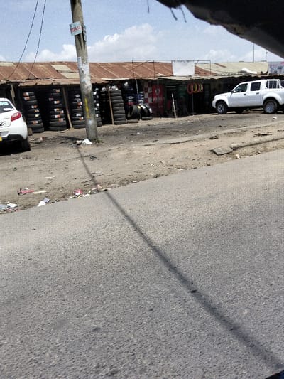 The image displays an outdoor street scene in Mkomani ward, Kenya, under a bright, partly cloudy sky. An asphalt road occupies the foreground, traversed by a long, diagonal shadow cast from a utility pole located slightly left of center. This pole has several posters affixed to its surface.
Adjacent to the road, a dirt and gravel shoulder stretches across the midground, covered with scattered litter including paper and plastic debris. Parked on this shoulder are two white vehicles: a hatchback, partially visible on the far left, and a four-door pickup truck positioned further right, facing left.
Behind the shoulder, a row of single-story commercial structures with corrugated metal roofs line the background. These are tire shops, characterized by numerous stacks of new and used tires displayed vertically and horizontally outside and within their open fronts. One shop features a red and white checkered facade with "AISAM AUTO..." and an "Airtel" logo visible. Another shop to its right has a sign indicating "GOODLUCK TYRES." One individual is seated outside the "AISAM AUTO" shop, to the right of the utility pole.