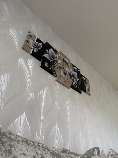 The image shows a wall with textured wallpaper in an interior setting. A set of five canvas prints depicting stylized floral designs in black, white, and beige are mounted on the wall in a linear arrangement. The wallpaper has a raised pattern that appears to be stylized feathers or leaves, with a subtle sheen. A portion of a textured upholstered furniture item, possibly a sofa, is visible in the foreground, filling the bottom of the frame. The lighting suggests an indoor environment, with a light source casting subtle shadows on the wall and artwork. The location of this scene is not specified in the image.