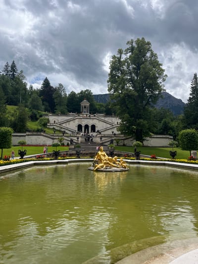 The image depicts the grand gardens of Linderhof Palace in Ettal, Germany. In the foreground, a large, greenish pond occupies the lower portion of the frame, with a golden sculpture of figures situated in its center. The sculpture appears to be a mythological scene with multiple human-like forms. The pond's edge is lined with a low, white wall, behind which a manicured lawn is visible, interspersed with flowerbeds displaying red and yellow blooms. Several black urns are strategically placed along the lawn.
Beyond the pond, a grand, multi-tiered staircase dominates the middle ground, leading up to a stately white building with arched entrances. At the very top of the staircase, a small, ornate pavilion with a dome is visible, housing a white statue. The architecture is classical in style. The building and terraces are surrounded by lush green grass and trees.
In the background, a dense forest covers a hillside, with a mountain range appearing in the far distance under a cloudy sky. The sky is filled with dramatic, dark gray clouds, suggesting overcast weather, with patches of lighter sky peeking through.
Several people are scattered throughout the scene. Two individuals are walking on the lawn in the middle ground, one in pink and the other in a striped top. Another person is visible on the steps of the building. In the foreground, on the far left, two people, possibly a couple, are standing near a black urn and trees. The overall impression is one of a majestic and well-maintained European palace garden, set against a natural mountainous backdrop.