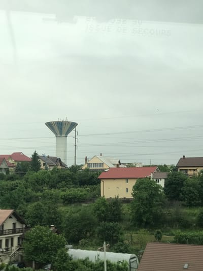 This image presents an exterior view of a residential area in the city of Suceava, Romania, captured on an overcast day from an elevated perspective, likely through a window, as indicated by faint reversed reflections of text ("29," "ISSUE DE SECOURS") in the upper sky.
The central and dominant feature is a tall, slender water tower with a white cylindrical base and an inverted conical tank atop. The tank's exterior is adorned with alternating vertical stripes of light blue and white, and various pieces of equipment are visible along its upper rim. To the immediate right of the water tower stands a tall utility pole supporting multiple horizontal power lines that traverse the mid-ground and upper portions of the scene.
Below the power lines and water tower, numerous residential houses are nestled amongst abundant green trees and dense vegetation, suggesting a hilly or undulating landscape. The houses exhibit varied architectural styles and roof colors, including prominent red, brown, and light brown/beige tiles. One house in the mid-ground, to the left of the water tower, features a red roof and light-colored walls. Another nearby house has a light brown roof with visible solar panels installed. Portions of other houses with brown roofs and white or beige facades are visible in the foreground and background.
The landscape is heavily forested with deciduous trees and bushes, providing a lush green backdrop to the residences. In the lower right foreground, a partial view of a brown-tiled roof is visible, while a white-covered greenhouse structure is partially seen in the lower left-center. The sky is uniformly light gray and overcast, diffusing the daylight. No people are visible in the scene.
