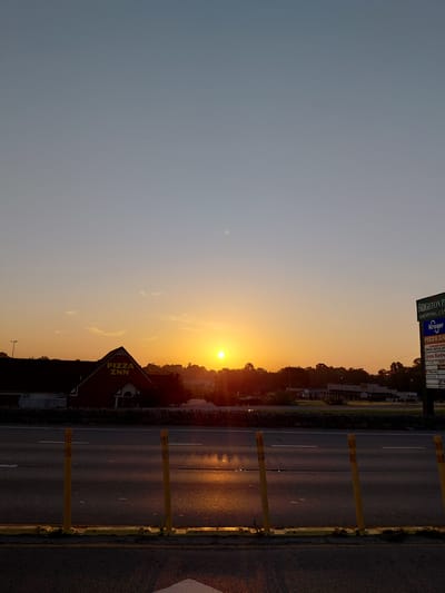 This is a landscape photograph taken at sunset in Frankfort, United States of America. The sun is setting over a tree line, casting a warm orange glow across the sky and reflecting on the wet asphalt of a road. The road is bordered by yellow posts and a stone wall. In the foreground, a building with a prominent "PIZZA INN" sign is visible, along with a separate sign indicating "BRIGHTON PARK SHOPPING CENTER" with other businesses listed. The scene suggests a commercial area at dusk. The sky transitions from a pale blue at the top to a vibrant orange near the horizon, indicating a clear evening. No people are visible in the image.