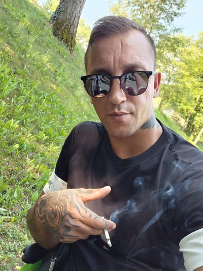 The image shows a man outdoors, holding a cigarette and smoking. He is wearing a black t-shirt and sunglasses. The man has tattoos on his hand and neck. The background is a grassy area with trees, suggesting a park or natural setting. The lighting indicates it is daytime, likely sunny. The image appears to be a selfie, with the man looking towards the camera. The scene is casual and relaxed. There is no visible text in the image. The orientation of the image is sideways.