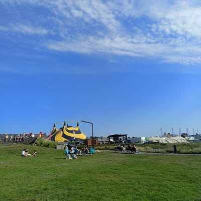 The image depicts a sunny outdoor scene in Ballybunion, Ireland, likely during a festival or event, with a clear blue sky dotted with wispy white clouds. In the background, a distinctive large tent with yellow and black stripes, characteristic of a circus or event structure, is prominently displayed. Adjacent to the tent, a stage area is set up with lighting rigs and speakers. Further to the right, several white marquees or tents suggest a larger gathering or market.
In the mid-ground, people are seen seated on a low stone wall, appearing to observe the scene or interact. Below them, on a grassy slope, a small group of people are relaxing on the grass, some sitting and talking, while others are reclining. To the right, several individuals are seated on park benches, also enjoying the outdoors. In the foreground, the expansive green grass covers the majority of the space, with a single seagull visible on the left. The overall impression is one of a leisurely day at an outdoor event.