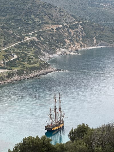A high-angle, aerial view of a scenic coastal landscape in Datça, Turkey, currently displayed with a 90-degree clockwise rotation. When viewed upright, the image features a three-masted sailing ship anchored near a rocky coastline. The ship, likely a tall ship or a replica, has a dark hull with distinctive yellow ornamentation along its upper deck. Its sails are furled, and intricate rigging is visible. On its stern, the word "EUROPA" is visible, along with a blue and white flag.
The ship rests in clear, calm turquoise water close to a rugged, rocky shore that transitions into a steep, vegetated hillside. The hillside is covered in dense green Mediterranean flora, with sparser trees closer to the water's edge. A narrow, winding road, possibly unpaved or lightly paved, traverses the hillside, following the contours of the land. Several utility poles are spaced along this road. A dark-colored car is visible on the road in the mid-ground.
The water surface shows ripples and reflections, indicating calm conditions under bright daylight. The overall impression is one of a serene and natural coastal environment, possibly a bay or cove, with a historic vessel as a central point of interest. There are no visible people or discernible activity on board the ship or along the coastline, suggesting a tranquil moment.