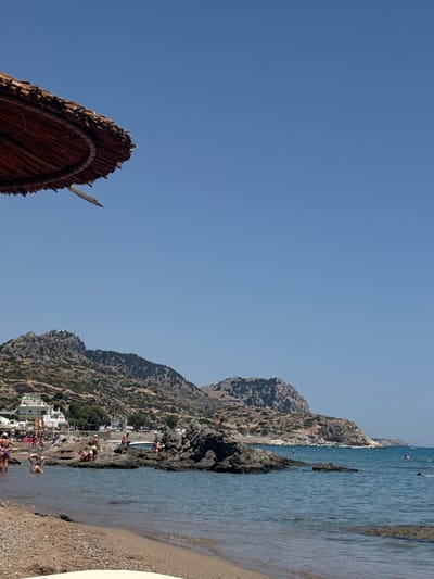 A wide-angle, eye-level shot captures a scenic view of a rocky beach in Archangelos, Greece. A thatched umbrella is in the upper left corner of the frame, with its edge casting a shadow. The azure sky fills the upper portion of the image.

In the mid-ground, a rocky shoreline meets the calm, clear turquoise water. Several people are visible in and around the water and on the rocks. Some are wading, while others are sunbathing or interacting on the rocks. A cluster of white buildings is nestled at the base of a rugged, mountainous landscape that dominates the background. The mountains are sparsely covered with scrub and trees. The beach itself is composed of sand and pebbles, with small waves lapping at the shore. The overall scene suggests a sunny, leisurely day at a coastal resort.