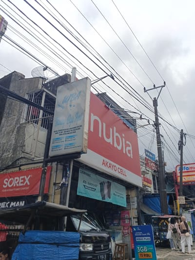 The image depicts a busy commercial street scene, presented as being in Palayan, Philippines. The sky is overcast, and numerous black overhead power and communication lines crisscross the upper portion of the frame.
Multiple multi-story buildings line the street. On the left, a concrete building with barred windows on an upper floor features a satellite dish on its roof. Below it, a white vertical sign advertises "WBA" with Indonesian text detailing the sale of various clothing and baby items. Adjacent to this, a red banner displays "SOREX".
Dominating the central mid-ground is a large red billboard with "nubia" in white lettering, and "AKAYO CELL" below it. Beneath this sign, a blue banner advertises a "SAMSUNG Galaxy A07" mobile phone. Further to the right, a blue and white "XL" banner promotes an internet package with Indonesian text, "Jaringan Internet Cepat XL BEBAS PUAS 3GB 3 hari 10 RIBU." A partially visible "Indomaret" sign is on the far right.
A dark-colored utility van, possibly a Suzuki Carry with a partially visible license plate "F 9045 TX," is parked on the left, partially obscured by a blue tarp and a makeshift stall structure. A wooden stool rests on the sidewalk in front of the "AKAYO CELL" establishment.
Three individuals, likely women wearing light-colored attire and head coverings, walk away from the camera on the sidewalk to the right. The top of a fourth person's head is visible in the bottom-left corner of the frame. Several utility poles with attached wires are scattered throughout the scene. The overall environment suggests a congested urban setting with various retail businesses.