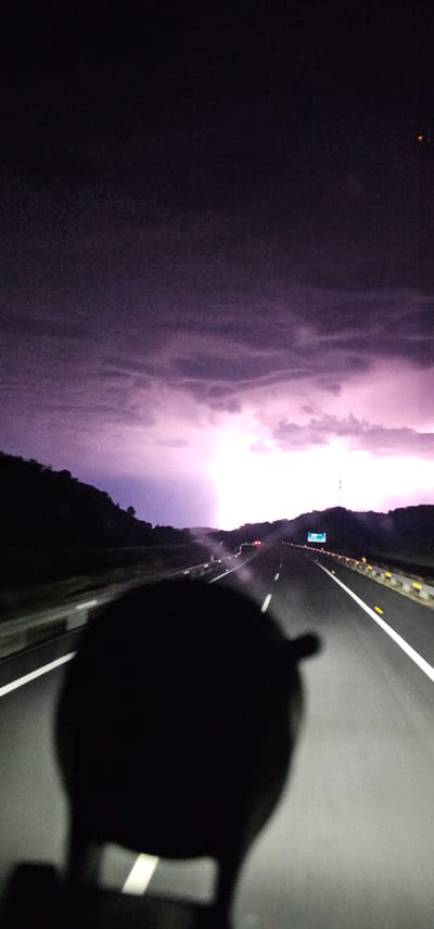 The image captures a highway scene at night, with a dramatic thunderstorm in the background. The sky is filled with dark purple and pink clouds, illuminated by flashes of lightning. The highway stretches into the distance, with white lines marking the lanes. On the right side of the highway, there is a guardrail with reflective markers. A dark, blurry object, likely a camera or a part of the vehicle from which the photo was taken, is in the foreground, obscuring the lower part of the image. Distant vehicles with red lights are visible on the highway. The landscape on the sides of the highway appears to be hilly and dark, suggesting a rural or semi-rural setting. The location is identified as Malpartida de Plasencia, Spain.