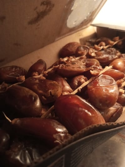 The scene depicts a close-up, upward-angled view of numerous dried dates contained within an open, corrugated cardboard box. The location is specified as B. Nam Ngao, Laos.
**Subjects/Objects:**
The primary subjects are a quantity of dates, numbering approximately 30-40 visible, varying in size and shape. They exhibit a deep brown to reddish-brown coloration, with some displaying a shinier, more intact skin surface while others appear slightly shriveled or textured. Several dates have small, fibrous, light-brown stem fragments or calyx structures still attached. Some dates show signs of splitting or minor damage.
The dates are situated inside a brown, corrugated cardboard container. The box appears partially open, with an upper flap lifted, exposing its interior surface which exhibits some minor smudges or discolorations. The lower right edge of the box's exterior is visible, showing faint printed text or markings, indicative of a product label.
**Environment/Lighting:**
The immediate environment is confined to the interior of the cardboard box. No external surroundings are clearly visible beyond the container. The lighting is dim and warm-toned, suggesting an indoor setting with artificial or low natural light. There are no visible individuals, actions, or interactions occurring within the frame.