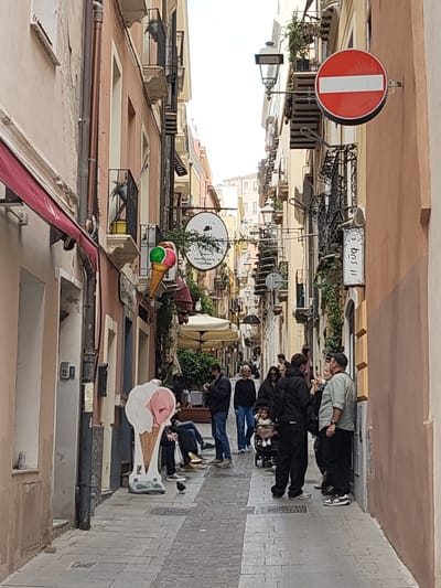 The image captures a narrow street scene in the city of Cagliari, Italy. The street is paved with grey stone bricks and lined with buildings of varying pastel colors (pink, peach, yellow) that are multiple stories high. Architectural details include small balconies with wrought iron railings, window boxes with plants, and awnings over storefronts. Several signs are visible along the street, including a large ice cream cone, a circular sign for a restaurant, and a "no entry" traffic sign.
A group of people is gathered along the street. Some are standing and talking, while others are seated. A woman is pushing a stroller. A life-size cutout of an ice cream cone stands at the street's edge. A small bird is on the pavement. The overall environment is bustling and suggests a tourist or commercial area.