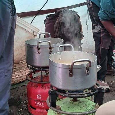 Within a light-colored canvas shelter at Gilman's Point, Tanzania, two large, silver-colored aluminum cooking pots are centrally positioned, actively heating. The pot in the foreground emits visible steam, indicating a light-colored liquid boiling within. Each pot rests on a black gas burner assembly, which is placed atop a gas cylinder. The mid-ground gas cylinder is red with "ORYX ENERGIES" branding, while the foreground cylinder is light green. Partial views of two individuals are present: on the left, a person wearing dark trousers and brown shoes stands near a woven basket. On the right, a person wearing dark and light-paneled trousers with a dark blue-green shirt is partially visible, with a dark-colored backpack leaning against the canvas wall behind the pots. The ground beneath the setup consists of exposed earth.