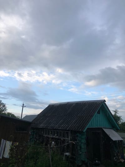 In the city of Staroe Istomino, Russia, an overcast sky looms above a rustic scene. In the foreground, a weathered wooden structure, possibly a shed or outbuilding, stands with its dark, corrugated roof slanting downwards. To its left, a smaller, darker wooden structure is visible, with laundry hanging on a line. A ladder is propped against the side of the larger building, suggesting recent activity or ongoing maintenance. A utility pole with power lines stretches across the middle ground, adding to the rural atmosphere. The overall impression is one of a quiet, utilitarian space, perhaps a backyard or a small farmstead, under a moody, cloudy sky.