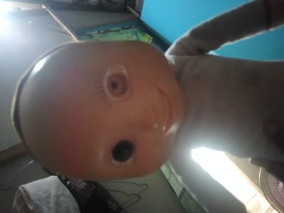 The image, taken from a low angle, shows a close-up of a doll's head and arm. The doll's face is mostly visible, with a round, orange plastic head featuring a painted eye with eyelashes and a mouth. A dark, circular opening, possibly a sound hole, is present below the eye. The doll's arm, made of a light-colored, fuzzy material, extends upwards and to the right, partially obscuring the background. The background is dimly lit and appears to be an indoor setting. Light streams in from the left, creating a bright glare across the scene. To the right, a portion of a fan is visible. The environment suggests a room, with wooden structures and a blue surface visible in the upper portion of the frame. The image does not provide any specific visual cues to pinpoint its exact location within Kedung Dowo, Indonesia, as it focuses solely on the doll and its immediate surroundings.