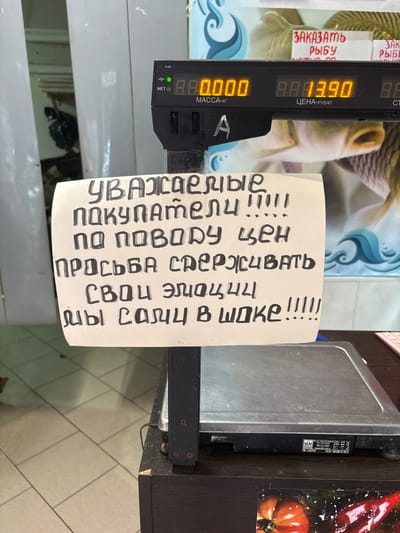 An electronic scale with a handwritten sign attached to its support post is depicted in a store. The scale displays numerical readings, and the sign is written in Russian in black marker on a piece of white paper. The sign reads, "Dear customers! Regarding the prices, please check your emotions, we are also in shock!!!!" In the background, there are images of fish and a price tag. The scale itself has labels and model numbers on its base. The environment appears to be a retail setting, possibly a fish market.