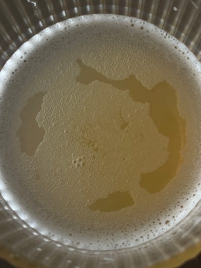 The image shows a close-up, top-down view of the foamy head on a beverage, likely beer, in a clear, ribbed glass. The foam is light yellow and uneven, with some larger bubbles and areas where the liquid underneath is visible, creating darker patches. The glass's textured rim is visible at the bottom and edges of the frame. The scene is illuminated by overhead light, casting soft shadows and highlighting the texture of the foam. The image is an abstract view of a drink and does not depict any specific location in Genemuiden, Netherlands.