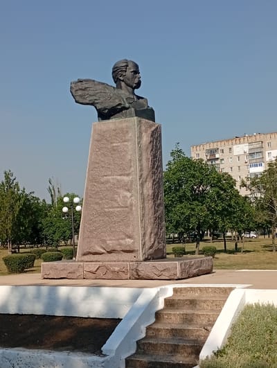 This media file depicts a bronze bust of a man on a large, granite pedestal. The bust shows the man from the shoulders up, with a stern expression and a mustache. His left arm is extended forward, appearing windswept or broken. The pedestal is tall and rectangular, with a rough, textured surface. At the base of the pedestal, there are several large, rough-cut granite blocks.
The statue is located outdoors in a park-like setting. Behind the statue, there are manicured green bushes, trees with lush green foliage, and a tall, light-colored apartment building with multiple balconies and windows. In the background, the sky is clear and blue, suggesting a sunny day. The light filtering through the trees and casting shadows on the white steps in the foreground indicate it is daytime.
The scene appears to be peaceful and static, with no discernible activity taking place other than the presence of the statue within its environment. The overall mood is one of solemnity and remembrance, characteristic of a public monument. No visible text is present in the image. The location context provided is Pokrov, Ukraine.