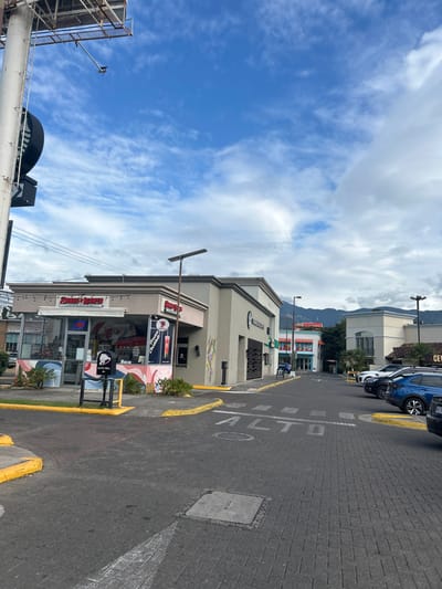 The image shows a street in Pozos, Costa Rica (latitude: 9.951, longitude: -84.194). The street is paved with grey brick and there is a sign on the ground that reads "ALTO," which means "stop" in Spanish. There are cars parked on the right side of the street. On the left side of the street is a commercial building with a "Subway" and "Pizza Hut" storefront. The building is beige and there are several green plants in front of the building. The sky is blue with white clouds. There are mountains in the distance. The image was taken during the day.