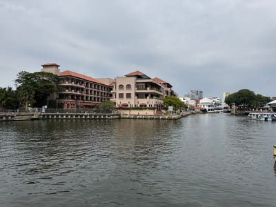 A waterfront scene features a river winding between buildings. On the left, a multi-story building with a red-tiled roof and balconies is nestled amidst trees. Along the riverbank, streetlights dot the path. Across the river, a mix of building styles is visible, with boats docked nearby. The sky is overcast. The river surface reflects the subdued daylight.
