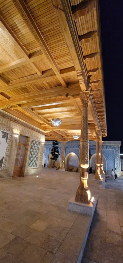 The image captures a nighttime scene outdoors at a location that appears to be a courtyard or walkway with traditional architecture. The scene is illuminated by artificial lighting, highlighting the intricate details of the structures.
The dominant subjects are the architectural elements, specifically a wooden roof supported by ornate columns and an adjacent building with decorative blue and white tilework. The wooden structure features a richly carved ceiling with repetitive geometric patterns and exposed beams. The columns are also carved with detailed designs and topped with intricate capitals. Hanging from the wooden ceiling are decorative lamps with a patterned, spherical design.
The walkway or courtyard is paved with light-colored stone tiles. In the background, the tiled building displays arched openings and windows with similar geometric patterns. There is a small tree planted in a pot within the courtyard.
There are a few people visible in the scene, appearing as small silhouettes in the distance, suggesting they are walking or standing in the courtyard. Their presence is minimal and does not detract from the architectural focus of the image.
The time of day is clearly night, indicated by the dark sky and the reliance on artificial lighting. There are no obvious cues about the weather, but the scene appears to be dry. The architectural style strongly suggests a location in Central Asia, and with the provided location context of Satkak, Uzbekistan, this is likely an area with a rich historical and cultural heritage. The overall mood is serene and possibly evokes a sense of wonder due to the detailed craftsmanship and ambient lighting. No visible text is present in the image.