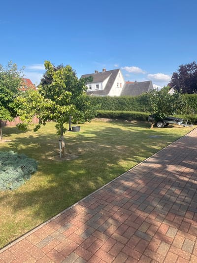 The image shows a residential backyard in Neustadt, Germany. The backyard features a paved brick walkway in the foreground, a well-maintained lawn, and several trees and bushes. In the background, there are houses with pitched roofs, some with visible windows and chimneys. A small trailer is parked on the grass near a hedge. The sky is blue with a few scattered white clouds. The scene appears to be a sunny day. There are no people visible in the image.
