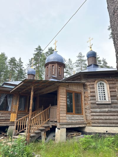 The image shows a wooden Orthodox church situated in a wooded area, likely in Nenyamyaki, Russia. The church is constructed from logs and features multiple domes topped with golden crosses. A covered porch with wooden stairs leads to the entrance on the left side of the building. The church has several windows, including a distinct arched window with a grid pattern on the right side. The surrounding environment is characterized by dense pine trees and lush green vegetation at the base of the structure. The sky is overcast, suggesting a cloudy day. There are no people visible in the image. The overall scene depicts a rustic and somewhat weathered church in a natural setting.