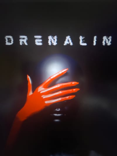 The image displays the word "DRENAALIN" in white, blocky, pixelated lettering across a dark, shadowy background. Below the text, a person's head is obscured by a dark, helmet-like object. A bright red, gloved hand with long, pointed fingernails is placed over the person's face, obscuring their eyes and nose. The hand appears to be a prominent focal point, with a glossy texture reflecting light. The overall mood is dark and somewhat mysterious. There is no information in the image to suggest a specific location in Punta Ala, Italy.