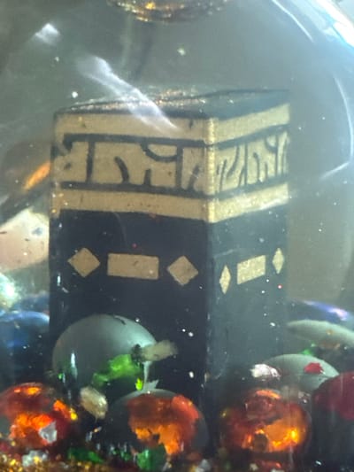 A black rectangular object with gold geometric designs sits amidst other colorful objects, possibly glass ornaments or beads. The designs include bands and geometric shapes. It appears to be submerged, possibly in a liquid or enclosed in a glass container, as there are visible distortions and reflections. The overall scene is somewhat blurry.
