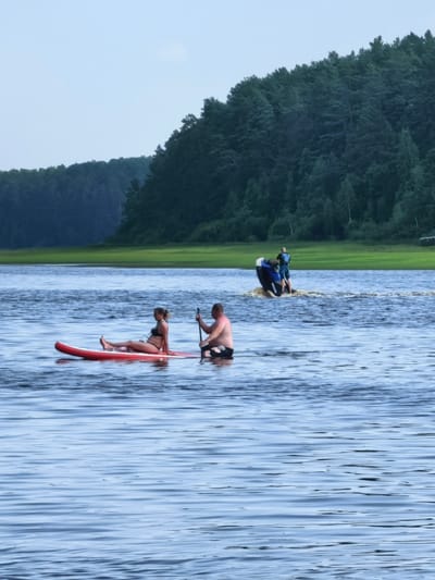 Two people are paddleboarding on a calm lake. In the foreground, a woman in a black bikini sits on a red paddleboard, facing left. A man in black swim trunks sits on a paddleboard behind her, facing right, holding a paddle. In the background, two people ride a jet ski, creating a wake. The water is blue and slightly rippled. Beyond the lake, a grassy area leads to a dense, dark green forest that covers a hillside. The sky is a pale blue with some hazy white clouds, suggesting it is daytime, possibly late afternoon or early evening. The overall scene is one of leisurely activity on the water in a natural setting. There is no visible text.