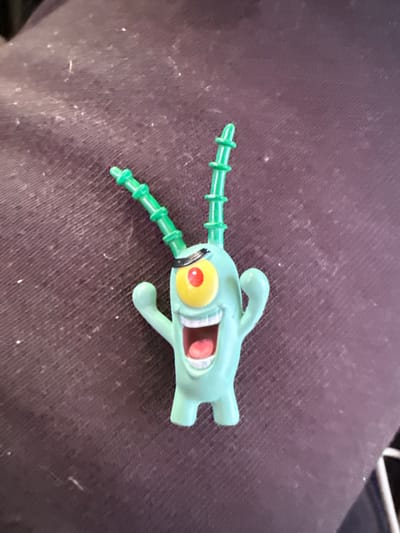 A light green plastic toy, resembling the character Plankton from SpongeBob SquarePants, is lying on a dark purple fabric surface. The toy has two green antennae with segmented details, a single large eye with a yellow iris and red pupil surrounded by a black band, and a wide open mouth revealing pink interior and white teeth. Its arms are outstretched. The fabric surface appears to be clothing, possibly pants or a jacket, and is covered in small white specks, likely lint or dust. The lighting suggests an indoor environment. No other subjects or discernible elements of a city or specific location are present.
