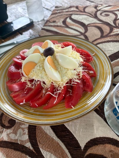 The central subject of the image is a colorful salad served in a round, clear glass plate with a distinctive yellow and white striped rim. The salad features thinly sliced red tomatoes arranged circularly around the edge of the plate. The interior of the salad is generously topped with shredded white cheese, likely Parmesan, and several halves of hard-boiled eggs, cut lengthwise to expose their white and orange yolks. A single dark, whole olive is placed near the center of the dish.

The plate rests on a textured tablecloth with an elaborate abstract pattern in shades of brown, beige, and white, featuring swirling and curvilinear designs. In the upper right corner, a clear glass is partially visible, bearing blue Cyrillic text, which appears to spell "БАЯ" or "БАЛ". In the lower-left, parts of a dark object, possibly a handbag or a wallet, and what might be cutlery handles are visible.

The scene depicts a prepared meal, suggesting a dining setting. The overall lighting indicates it is likely daytime or a brightly lit indoor environment. The image is taken from an overhead perspective, showcasing the arrangement of the food. The location context provided is Sushina, Bulgaria, which aligns with the Cyrillic script visible on the glass.