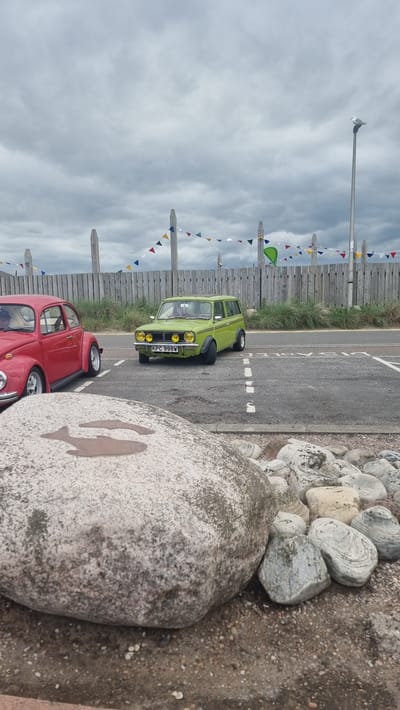 The image shows a roadside scene near Montrose, United Kingdom. In the foreground, a large, light-colored boulder is prominent, with a dark silhouette of two fish painted on its surface. To the right of the boulder is a gravelly ground.

Beyond the boulder and gravel, a road is visible, marked with white lines. A lime green car, possibly a classic Mini variant, is parked on the road, facing forward. It has a license plate reading "KPC 998W". Behind it, partially visible, is a red car, appearing to be a classic Volkswagen Beetle.

A wooden fence runs along the left side of the road, adorned with a string of colorful triangular flags. Beyond the fence, there is green grass and some vegetation. In the distance, a lamppost stands tall against a cloudy sky. The overall atmosphere suggests an overcast day, possibly late afternoon or early morning due to the diffused light. The presence of the flags and the beach-like setting (indicated by the rocks and gravel) suggests a coastal location, likely a holiday or recreational area. There are no people visible in the image.
