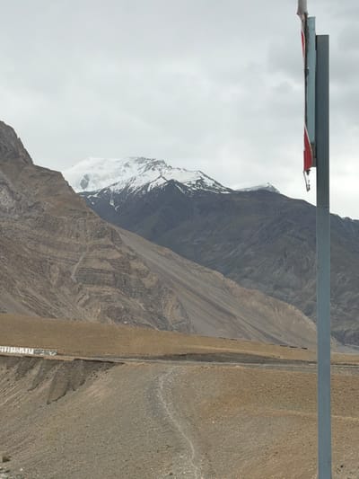 A vertical, full-shot image depicts a mountainous landscape under a cloudy sky. In the foreground, a tall, gray pole with a partially visible red and white sign is positioned on the right side. The terrain slopes upwards from the foreground to midground, featuring dry, rocky ground and a winding dirt road. A low, white structure with a patterned camouflage-like front runs along the edge of the road in the midground. Beyond this, imposing, layered mountains rise, their sides textured with erosion. The highest peaks in the background are capped with snow and partially obscured by clouds. The scene is situated in Zangla, India, a remote village in the Himalayas known for its stark, beautiful terrain.