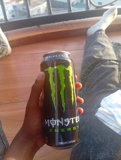 The image depicts a close-up, elevated view of an individual holding a can of Monster Energy drink. The person, whose dark-skinned right hand holds the black can with its prominent green "M" logo and "ENERGY" text, is seated on a light brown surface, likely wood planks or tiles. The top of the can indicates "TAURINE + GINSENG" and the bottom "CONTAINS CAFFEINE". The individual wears grey denim jeans; one leg is visible, with a foot clad in a white sock. To the right of the person's leg, a stack of folded white and blue checkered fabric, possibly napkins or a small towel, rests on the surface. In the background, beyond a dark metal railing, an urban environment is visible under natural light, indicative of the city of Murang'a, Kenya. The distant cityscape appears somewhat blurred, showing buildings with light-colored roofs and what appears to be a street or open area. No specific landmarks within Murang'a are discernible from this perspective.