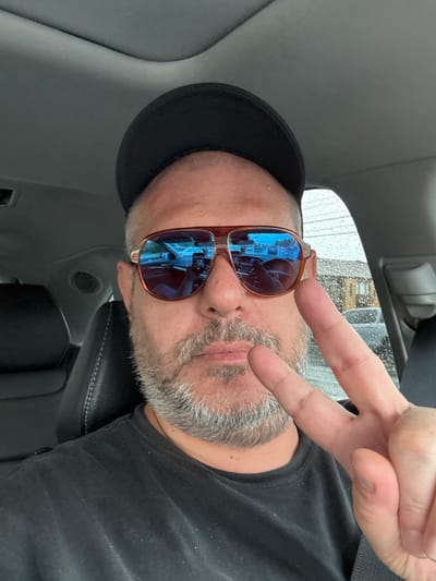 A man in a black t-shirt and a black baseball cap, wearing aviator sunglasses, takes a selfie in the driver's seat of a car. The man has gray facial hair and is holding up two fingers in a peace sign. The car's interior is visible, including black leather seats and a car ceiling. Outside the car window, there are raindrops and a residential area with houses and cars. The scene does not provide enough visual information to determine the specific location within Philadelphia, United States of America.