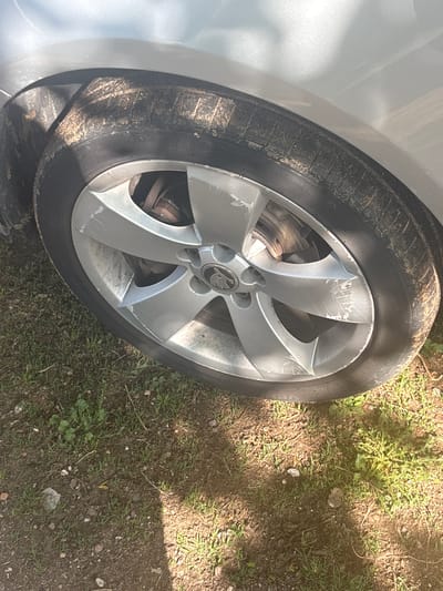 The image shows the front driver's side wheel of a silver Škoda car. The wheel rim has a five-spoke design with the Škoda logo in the center. The rim is scratched and dirty. The tire is also dirty and shows signs of wear. The car is parked on a patch of dirt and grass. The location is likely in Oriahovo, Bulgaria. No people are visible.