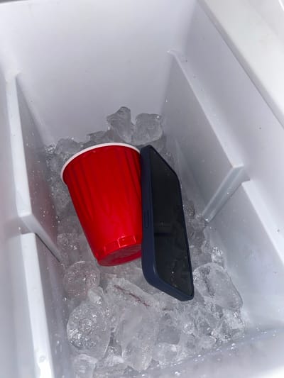The image shows the inside of a white cooler filled with ice. A red plastic cup is partially submerged in the ice, and a black smartphone rests on top of the cup. The background is white, indicating the interior of the cooler. The image contains no people, actions, or interactions. There is no information available in the image to support that the image was taken in Santa Cruz Tehuixpango, Mexico.