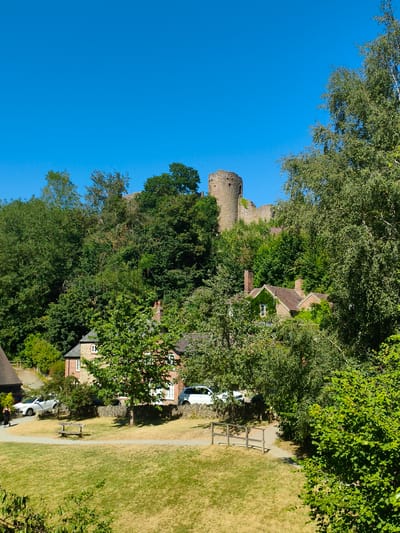 The image depicts a sunny day in Ludlow, United Kingdom, featuring a view of Ludlow Castle ruins atop a tree-covered hill. In the foreground, a grassy field leads to a pathway with a wooden bench. Adjacent to the pathway are several parked cars, including a white SUV and a white sedan. Behind the cars, a stone wall separates the road from a cluster of houses. One house, partially covered in ivy, has a visible window and a chimney. Another brick house is visible to its right. A person in dark clothing is walking on the pathway. The sky is clear blue. The overall scene conveys a tranquil, rural atmosphere with a historical landmark as a backdrop.