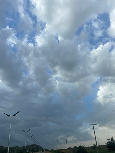 The image depicts a road in Barakin Kuru III, Nigeria, under a dramatic, cloudy sky. A line of streetlights runs along the left side of the road, with several poles of varying heights casting long shadows. On the right side of the road, utility poles with wires stretch into the distance. The landscape is characterized by rolling hills and sparse vegetation. The overall atmosphere is somewhat overcast, with patches of blue sky visible through the dense clouds. A few vehicles can be seen on the road, suggesting ongoing traffic. The scene appears to be a typical rural or semi-urban setting in Nigeria, with infrastructure for lighting and power.