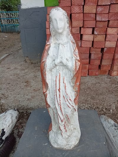 This image features a weathered statue of the Virgin Mary, predominantly white with faded reddish-brown and gold accents. The statue is depicted in a prayerful pose, with hands clasped at chest level. It stands on a grey stone slab, which is itself placed on a rough, gravelly ground. The statue's surface shows signs of age and deterioration, with paint peeling and chipping.
In the background, to the left of the statue, is a grey painted wall and a blue metal fence with decorative scrollwork, hinting at a yard or garden area. Beyond the fence, greenery is visible. To the right of the statue, a stack of red bricks is piled high, their textured surfaces clearly defined. The overall setting appears to be outdoors, possibly a construction site, a garden, or a religious compound, given the presence of the statue and building materials. The lighting suggests it is daytime, with no specific indications of weather conditions. There is no visible text in the image. The focus is entirely on the solitary statue and its immediate surroundings.