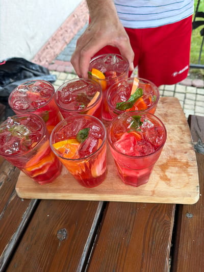 A person's hand is reaching for one of seven glasses filled with a red beverage and ice, garnished with citrus slices and mint leaves. The glasses are arranged on a wooden cutting board which rests on a dark wooden table. The background shows a white wall, a red checkered pattern, and some greenery, suggesting an outdoor setting.