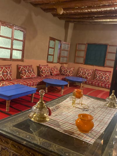 The image depicts an indoor seating area within a dwelling in Douar Ait Daoud, Morocco. The room features a rustic design with mud-plastered walls and a thatched roof supported by wooden beams.
Seating arrangements consist of a long, cushioned bench along the wall, adorned with red and white patterned cushions. In front of the bench are two low, bright blue tables. The floor is covered with a red and black striped carpet.
On a dark, intricately patterned table in the foreground, several decorative items are placed. These include a golden ornate stand holding a small orange pot, and another golden decorative piece with a red tassel. A white cloth with a red and blue checkered pattern is spread on this table.
Several windows with wooden frames are visible in the background, some with textured glass and others with dark green shutters. The overall impression is of a traditional, cozy living space. There are no people visible in the image.
