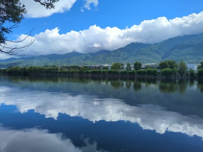 This is a landscape photograph taken from the edge of a body of water, likely a lake or calm river, with a reflection of the sky and surrounding scenery. The scene depicts a serene natural environment with a focus on the interplay between the sky, mountains, and water.

In the foreground, the dark, still water occupies the lower half of the image, acting as a mirror. It reflects the bright blue sky, fluffy white clouds, and the lush green mountains in the background. The reflections are distorted by gentle ripples on the water's surface, creating abstract patterns of light and color.

Beyond the water, a line of trees with full, green foliage forms a dense border, suggesting a park or a vegetated shoreline. Behind the trees, a range of verdant, mist-shrouded mountains rises, their slopes covered in dense vegetation. Some of the higher peaks are partially obscured by clouds, adding a sense of depth and mystery to the landscape. A small, scattered settlement or town can be glimpsed nestled at the foot of the mountains.

The upper portion of the image is dominated by a bright blue sky filled with dramatic, voluminous white clouds. A few bare branches from a tree in the upper left corner frame the sky, adding a touch of foreground detail. The lighting suggests it is daytime, possibly late morning or early afternoon, with bright sunshine illuminating the scene. The overall mood is peaceful and tranquil, evoking a sense of natural beauty and calm. There is no visible text in the image.