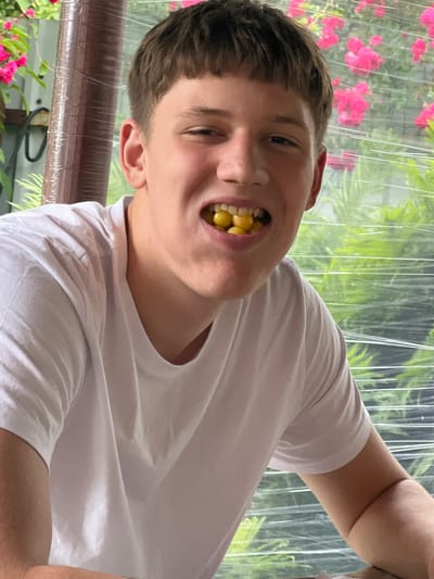 A young man with short brown hair and a white t-shirt is holding several yellow gumballs in his mouth. He is smiling broadly, with his teeth and the gumballs visible. The background is blurred, showing a window with pink flowers and green foliage visible outside. The image appears to be taken outdoors, possibly in a residential area. The exact location in Андрэеўшчынскі сельскі Савет, Belarus cannot be determined from the image alone.