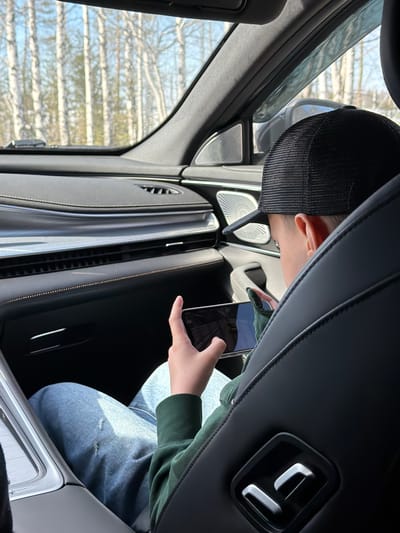 An individual is depicted from a rear-angle perspective, occupying the front passenger seat of a vehicle. The person wears a black mesh baseball cap, a dark green long-sleeved shirt, and light blue jeans with a small tear on the left thigh. Their left hand, partially covered by a green glove, holds a black smartphone horizontally, with its screen oriented towards the viewer's right, suggesting active engagement. The vehicle's interior features black leather upholstery with visible stitching on the seats and dashboard. Silver-colored, geometric seat adjustment controls are located on the side of the passenger seat. The dashboard displays horizontal air vents, a textured silver trim accent, and fine dotted stitching in a light contrasting color. A circular speaker grille is visible on the passenger side door panel. Through the vehicle's front windshield, a blurred exterior scene reveals numerous white-barked trees, consistent with birch, beneath a clear blue sky, indicating a wooded or forested environment. The specific location in Bolgury, Russia, cannot be independently confirmed from the visual information presented.